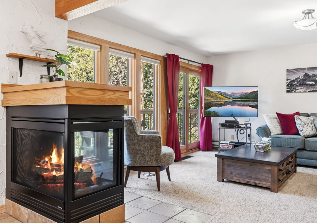 Curl up by the crackling fireplace as natural light floods through expansive windows framing mountain views, inviting you to unwind in this cozy living room.