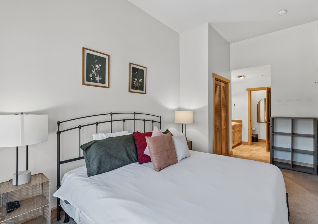 Your restful bedroom awaits with crisp white linens accented by jewel-toned pillows, a sleek black metal headboard, and soft natural light that creates a calming, serene atmosphere.