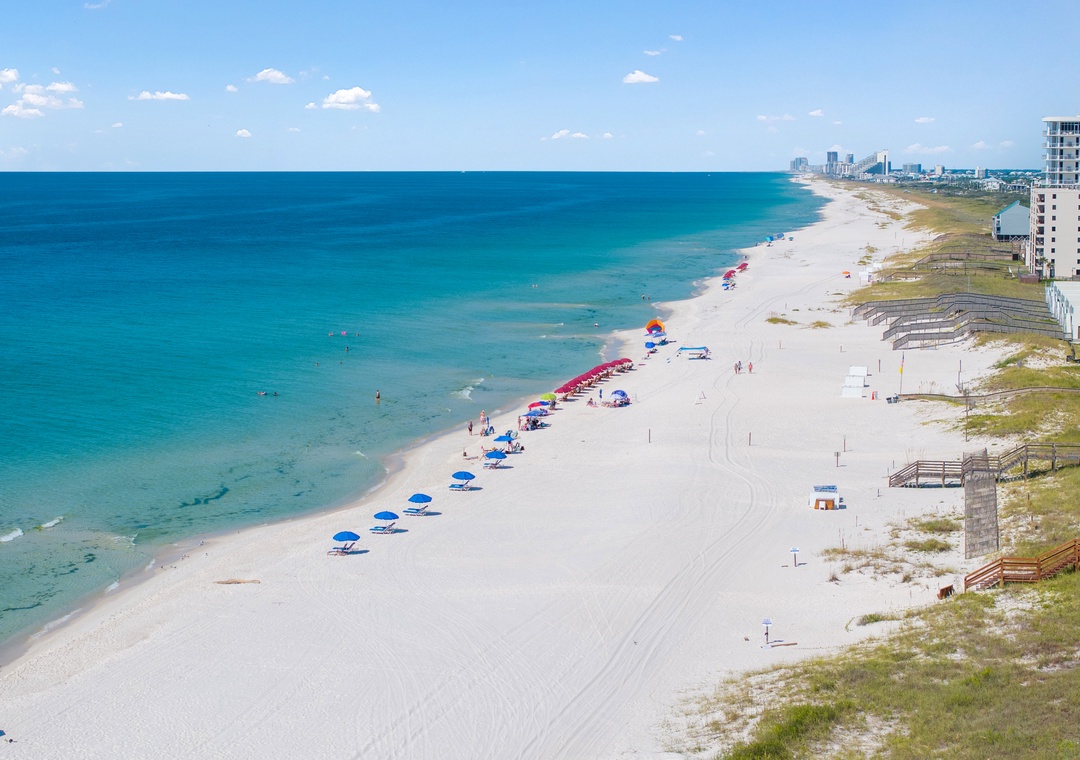 An elevated view showcases pristine white sands meeting turquoise waters along a stunning coastline, with colorful beach umbrellas dotting the shore and coastal buildings framing the horizon.