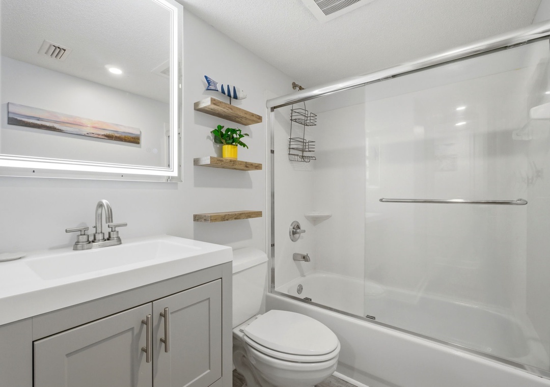 Freshen up in your bright, spotless bathroom featuring a tub-shower combo, sleek vanity, and thoughtful touches like floating wood shelves and charming coastal décor—perfect for your daily routine.
