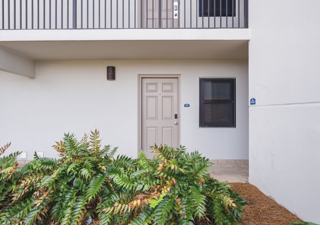Ocean Breeze W 105- The facade shows a clean exterior with a welcoming entrance door beneath a balcony, surrounded by vibrant landscaping with ferns in mulched beds.