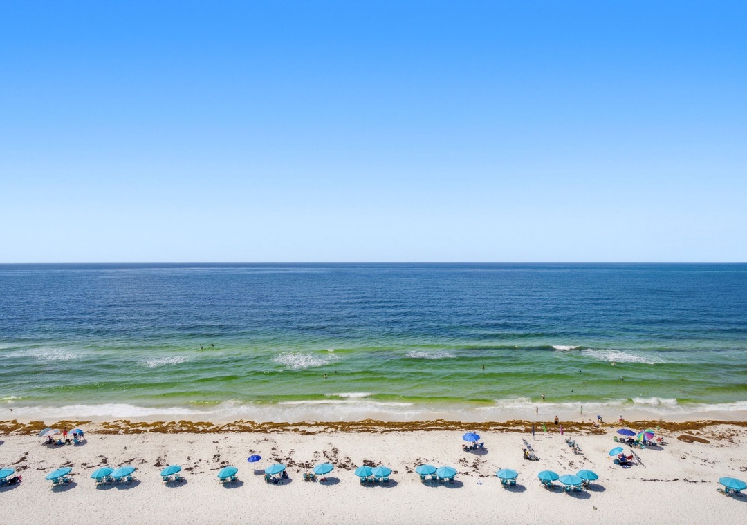 Aerial view showcasing the pristine beachfront location with turquoise waters, white sand, and rows of complimentary beach chairs and umbrellas for guests.
