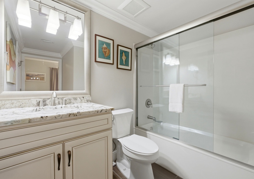 Step into this pristine bathroom with elegant marble countertops, a spacious glass-enclosed tub-shower, and coastal-inspired artwork that creates a serene retreat for your morning routine.