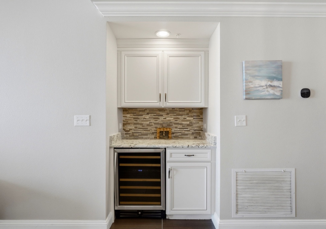Enjoy coastal elegance in your private wet bar, featuring sleek white cabinetry, granite countertops, a stylish mosaic backsplash, and a chilled wine fridge ready for your evening drinks.