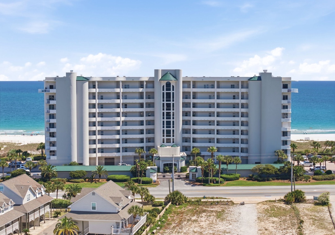 A beachfront condominium building situated along a pristine stretch of white sand beach, with tropical landscaping and neighboring residential properties creating a welcoming coastal community.