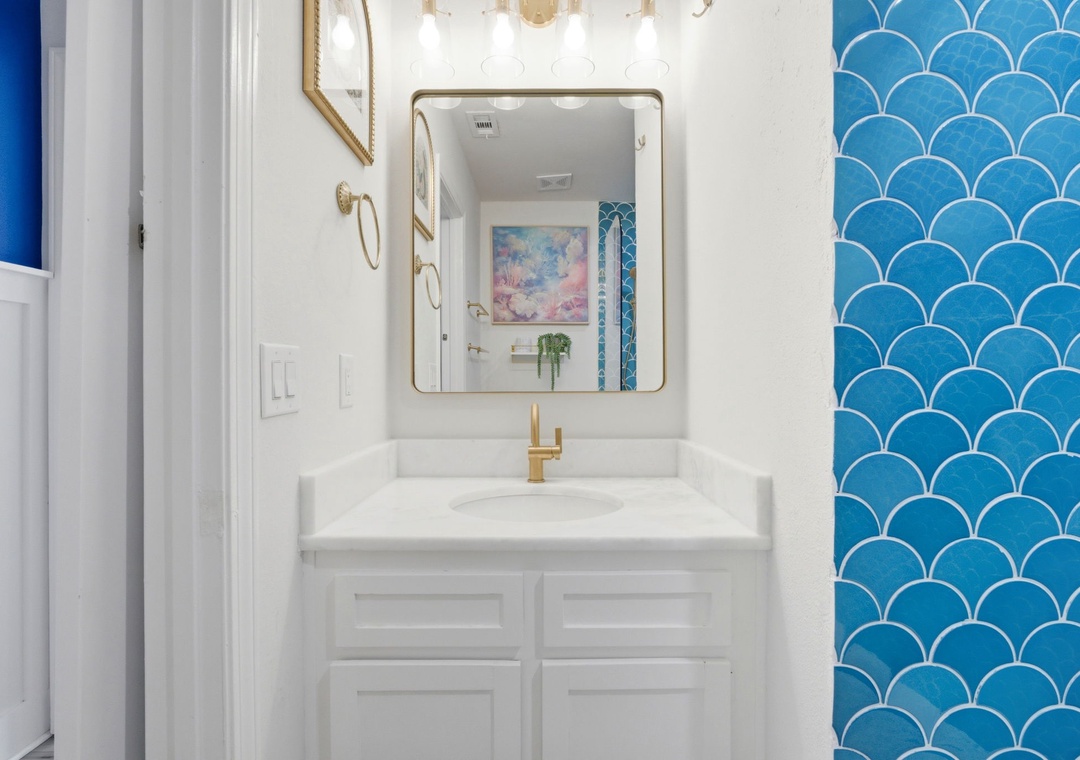 North Lost Key Landing B2- Refresh in this bright bathroom featuring a gleaming white vanity with gold fixtures, chic scalloped tile, and a golden-framed mirror that reflects coastal-inspired art.