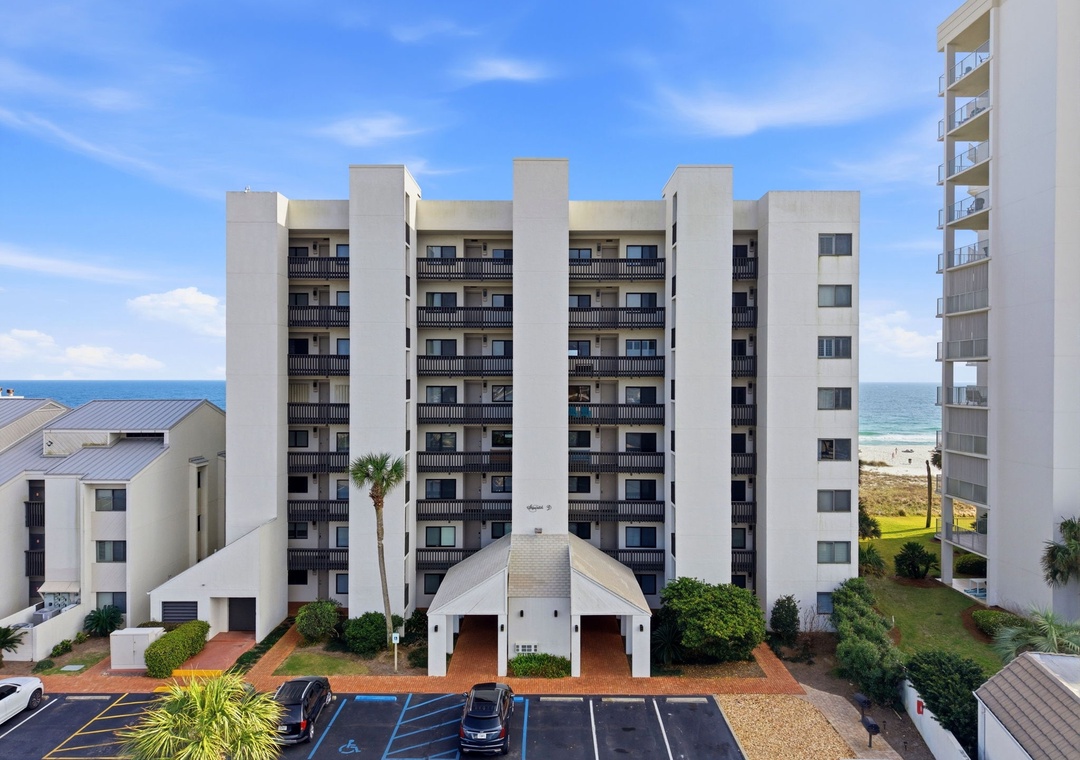 Shipwatch D803- A mid-rise beachfront property featuring multiple balconies and modern design, situated steps from the Gulf of Mexico. The building is surrounded by tropical landscaping and offers direct beach access.