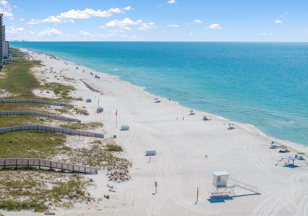 Pristine white-sand beach with crystal-clear Gulf waters stretching endlessly, captured from an elevated perspective showing the property's stunning beachfront location.
