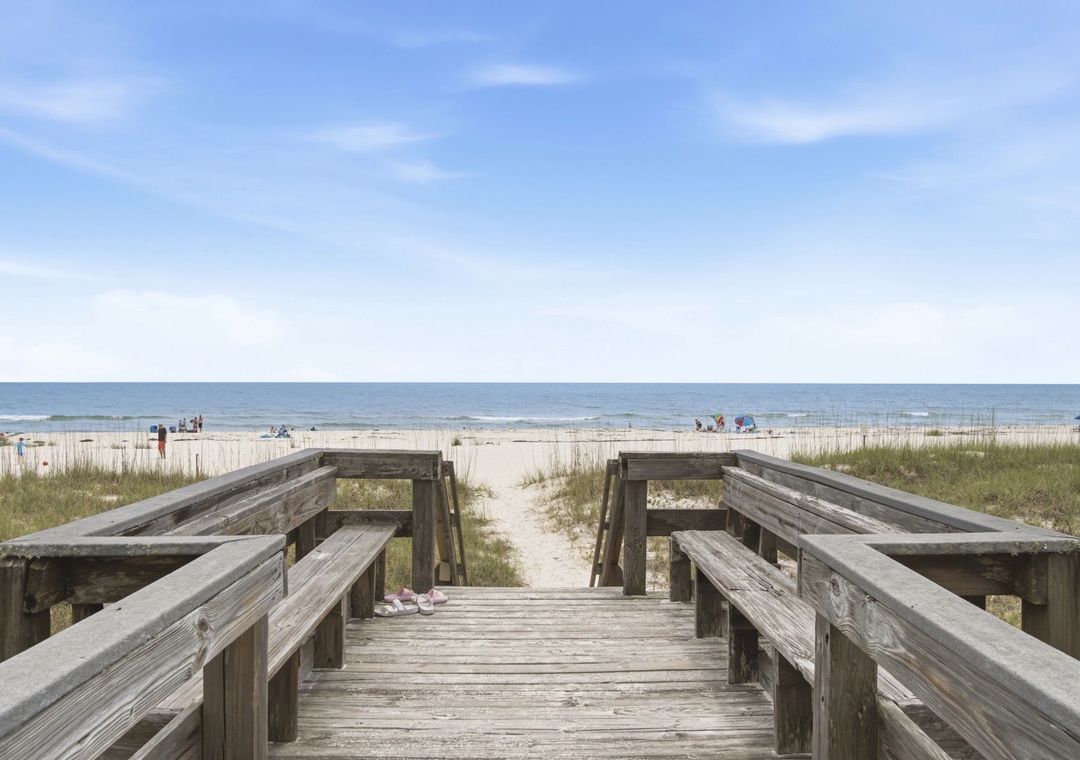 A wooden boardwalk leads across coastal dunes to a pristine sandy beach, with calm blue waters stretching to the horizon under a clear sky.