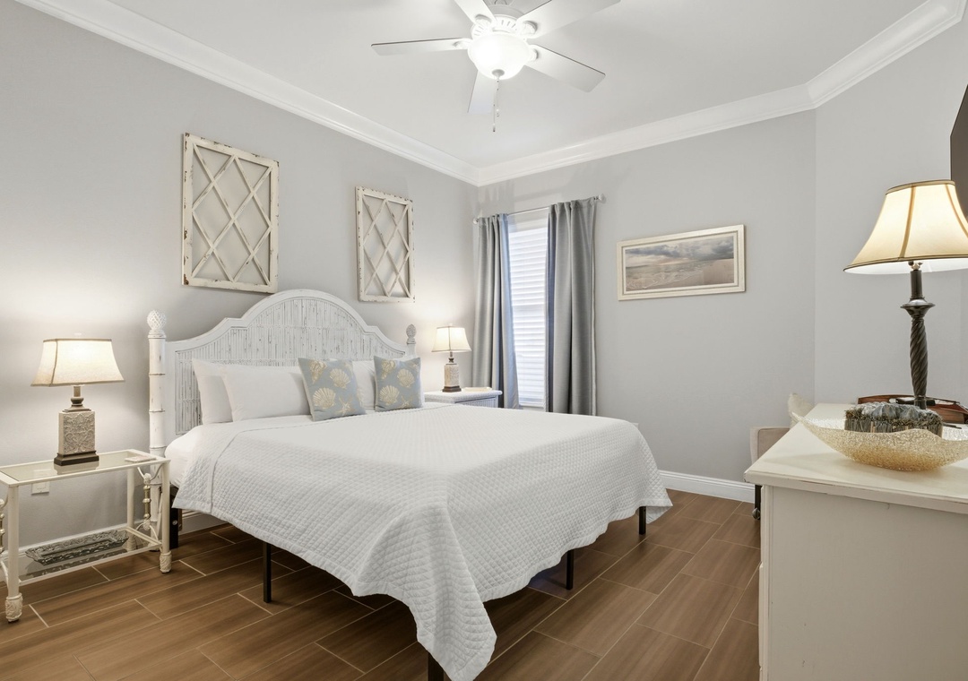Your serene retreat features a plush bed with crisp linens, coastal décor, and gentle ceiling fan breezes. Soft lighting and warm wood floors invite you to unwind in comfort.