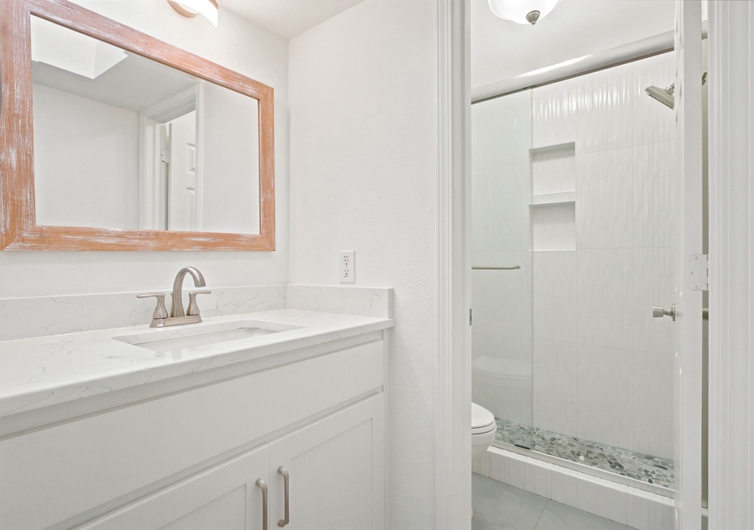 Gulfside Villas 2B- Your bathroom features a modern vanity with marble countertop, warm wood-framed mirror, and a spacious walk-in shower with built-in shelving for your convenience.