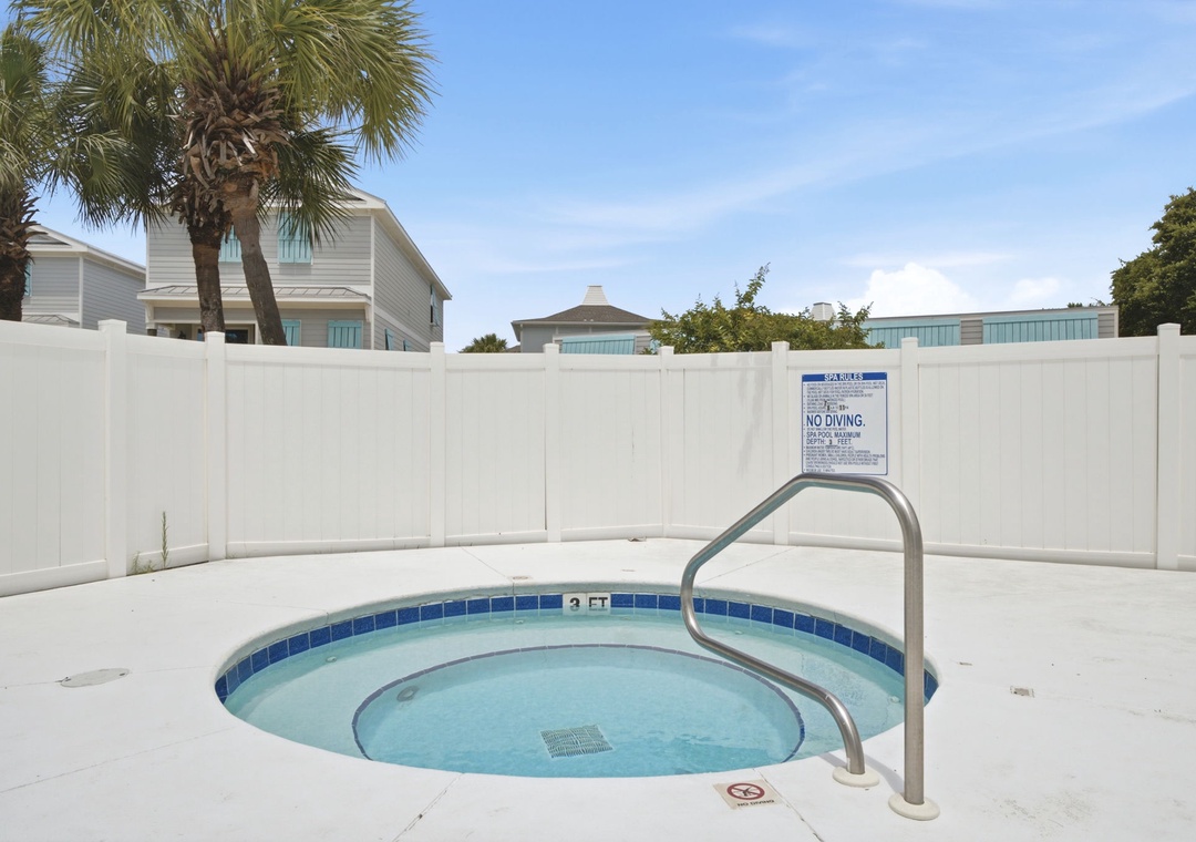 A private hot tub surrounded by clean white fencing, nestled among palm trees with spacious vacation homes in the background. Perfect for unwinding after a day at the beach.