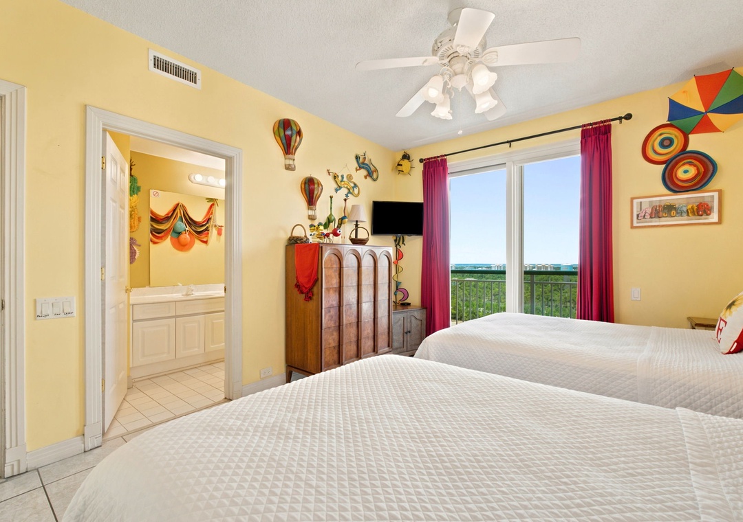 Beach Colony W13A- Step into your cheerful, sun-filled bedroom with two plush beds, whimsical décor, and a private balcony overlooking greenery and distant waters—your perfect retreat awaits.