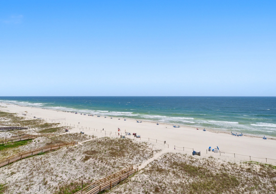 Shipwatch D803- Elevated coastal view showcasing pristine white sand beach stretching along emerald Gulf waters, with boardwalk access and beachgoers dotting the shore under clear skies.