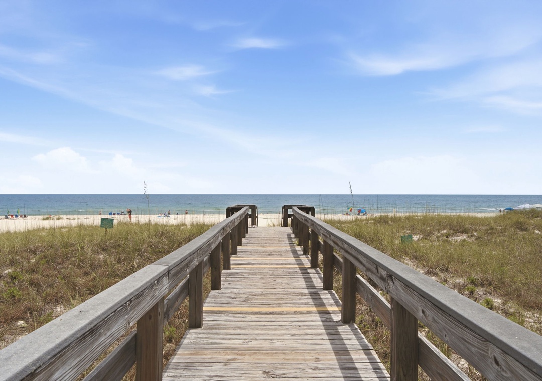 A wooden boardwalk leads through coastal dunes to pristine sandy shores, where calm waters meet clear blue skies—an inviting path to your private beach escape.