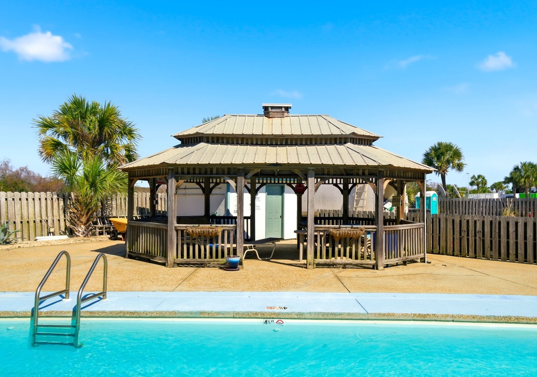A covered gazebo with poolside seating and palm trees creates a relaxing area for the property, located within a tropical landscape under clear blue skies.