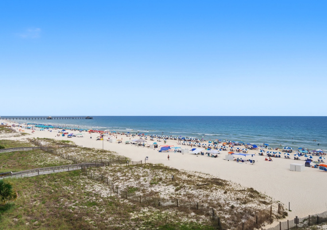Wide sandy beach stretches along clear blue waters, with gentle waves and a fishing pier extending into the horizon, marking the coastal setting of the property.