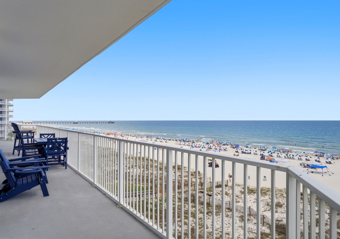 This spacious balcony offers stunning beachfront views with comfortable seating for relaxation. The white sand beach stretches along the sparkling turquoise waters, with a fishing pier visible in the distance.