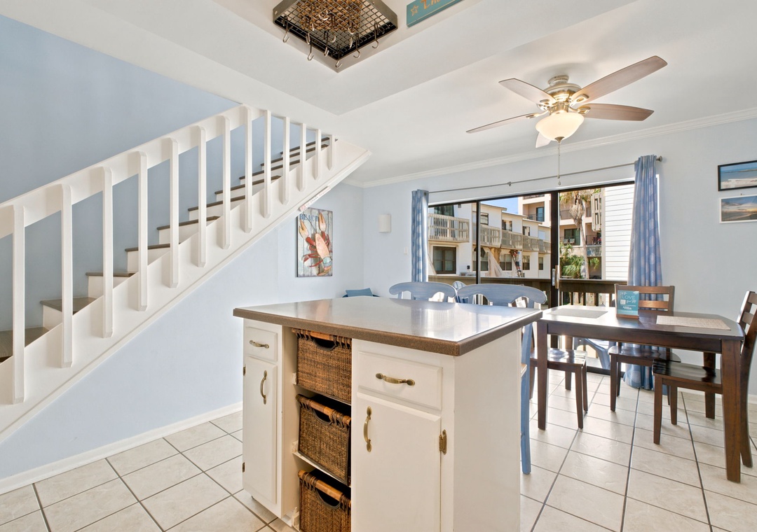 Picture yourself cooking and dining in this bright, airy space with coastal charm. Soft blue walls, a kitchen island with woven baskets, and a welcoming dining table set the scene for easy beachside living.