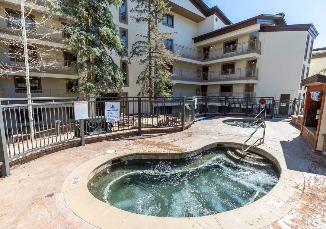 The property exterior featuring multi-story residential buildings surrounding a courtyard area with hot tubs and mature evergreen trees.