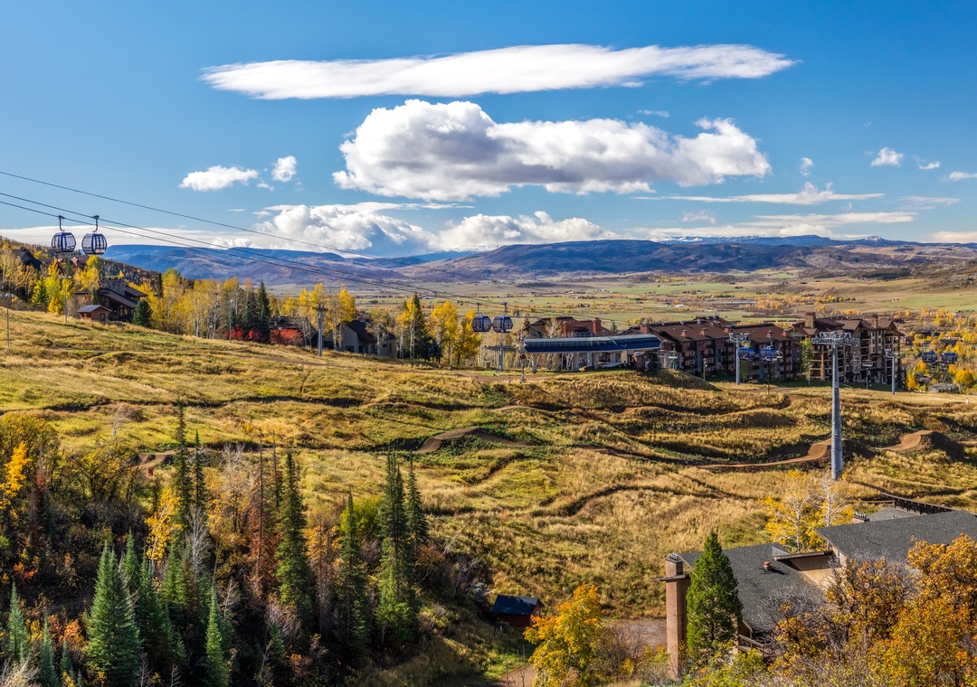 Mountain resort area with sweeping views across golden autumn valleys, rustic lodges, and distant ski lifts under a brilliant blue sky.