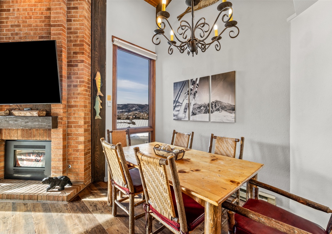 Gather around your rustic dining table where mountain artwork meets cozy warmth from the brick fireplace and elegant chandelier above.