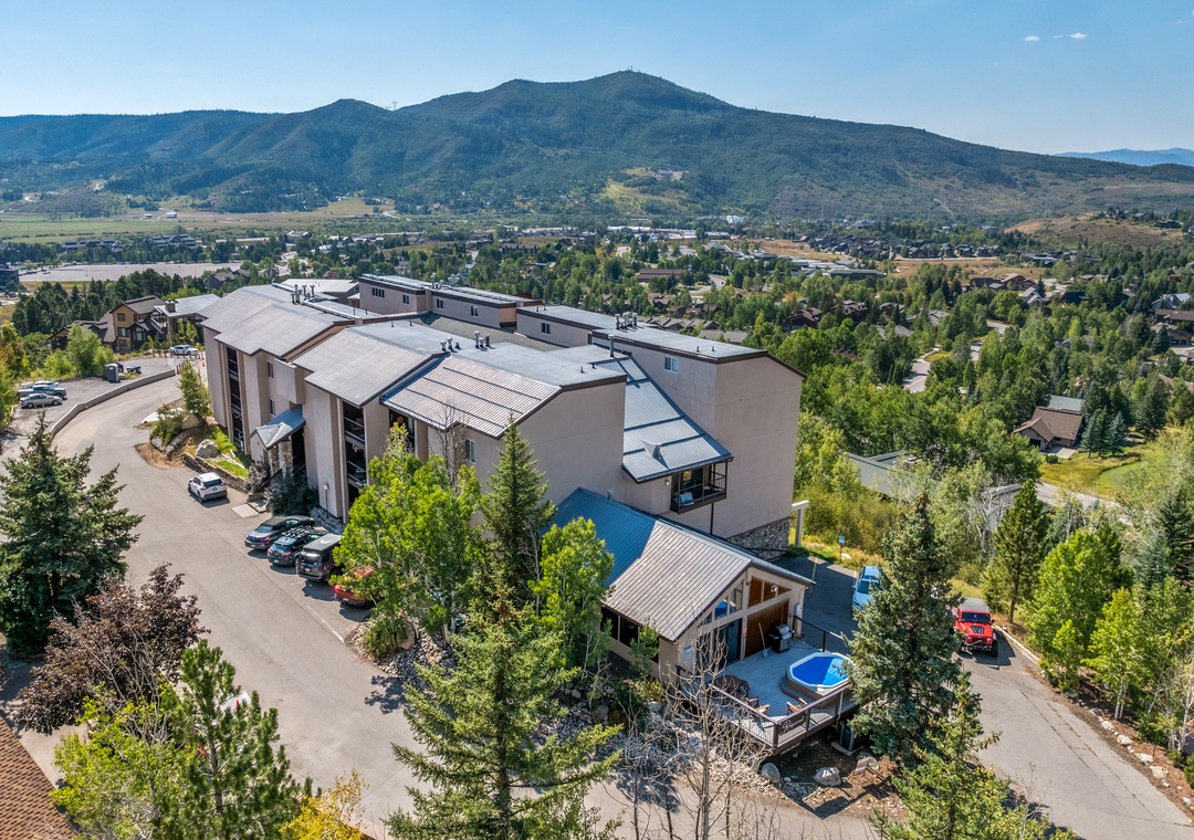 Aerial view of the property nestled among evergreens with mountain terrain in the background and nearby neighborhood visible below.