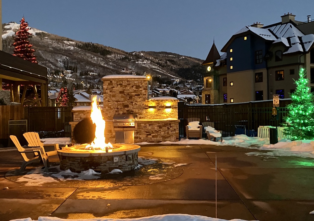 A roaring firepit and outdoor BBQ create a warm gathering spot under snowy mountain skies, perfect for alpine evenings with s'mores and stories.