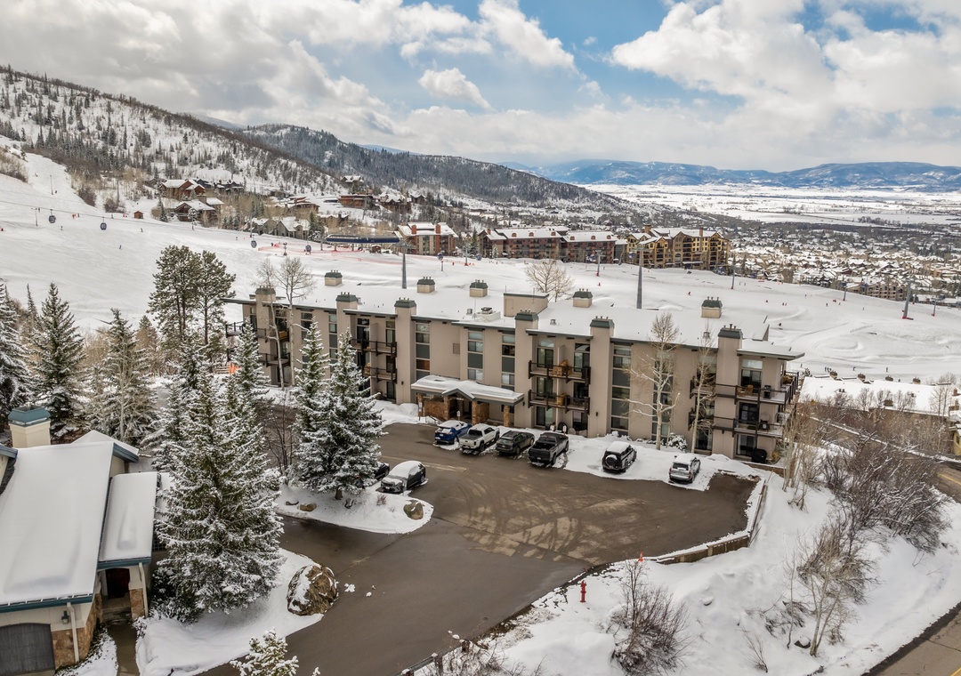 This mountain resort sits amid snow-covered slopes, with ski lifts visible just beyond the property and panoramic views of the valley below. The building is surrounded by evergreen trees and offers direct ski-in/ski-out convenience.