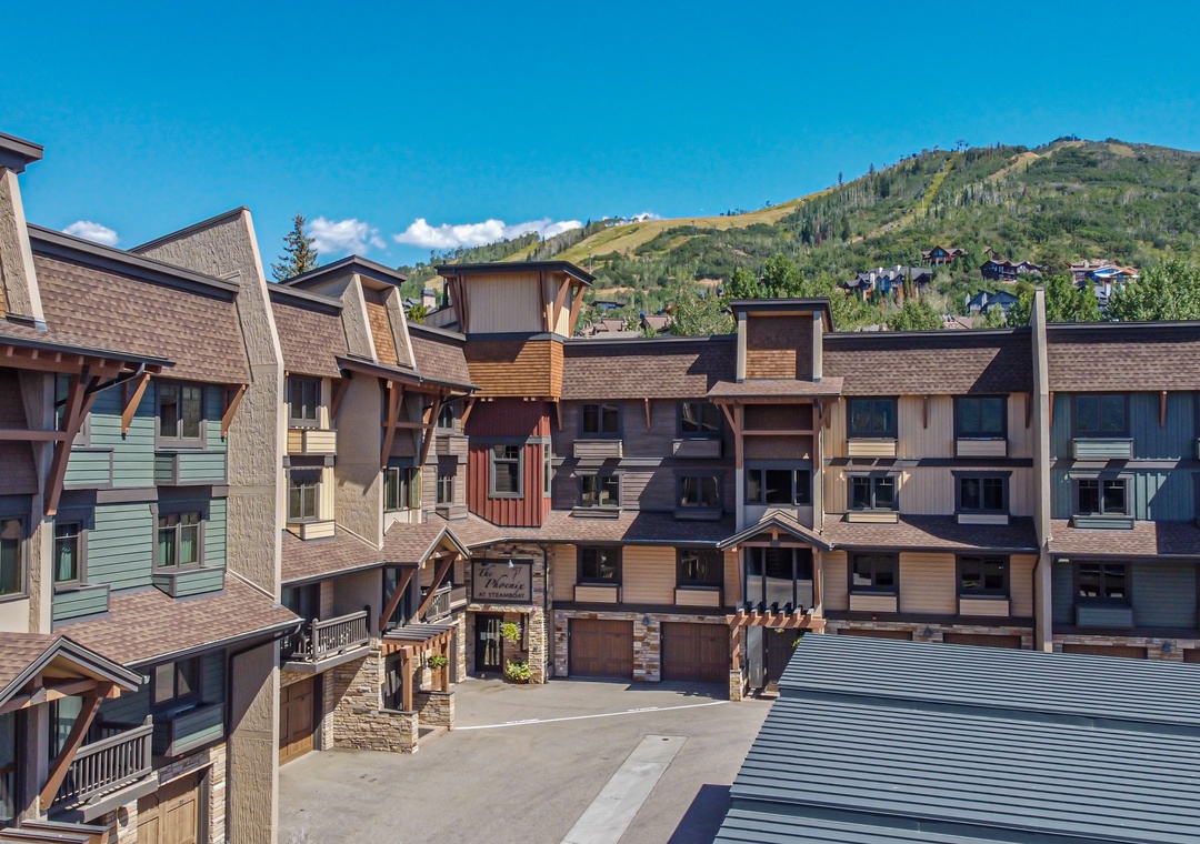 Multi-level condominium complex set against tree-covered slopes under clear summer skies, with convenient parking and mountain access nearby.