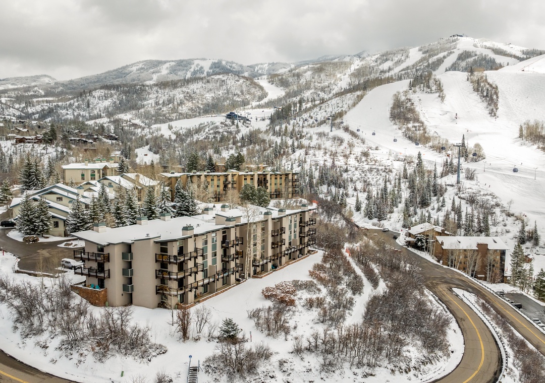 An aerial winter view showcasing the property nestled at the base of snow-covered ski slopes, with mountain terrain and ski runs visible in the surrounding landscape.