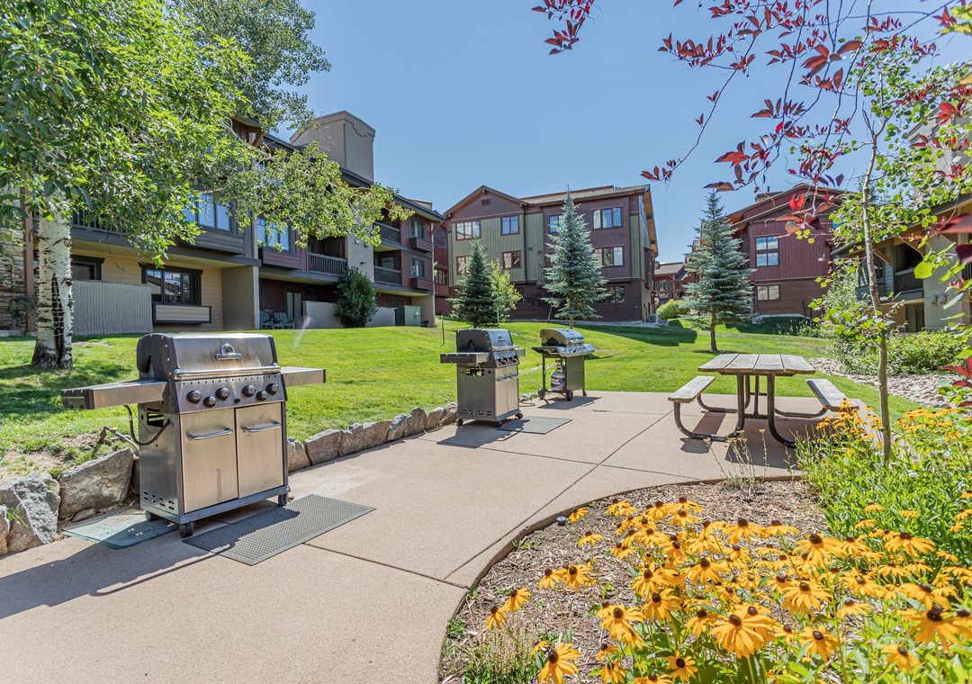 A sunny shared courtyard featuring multiple gas grills and picnic seating, surrounded by mature trees, colorful gardens, and conifer landscaping—perfect for enjoying the mountain air.