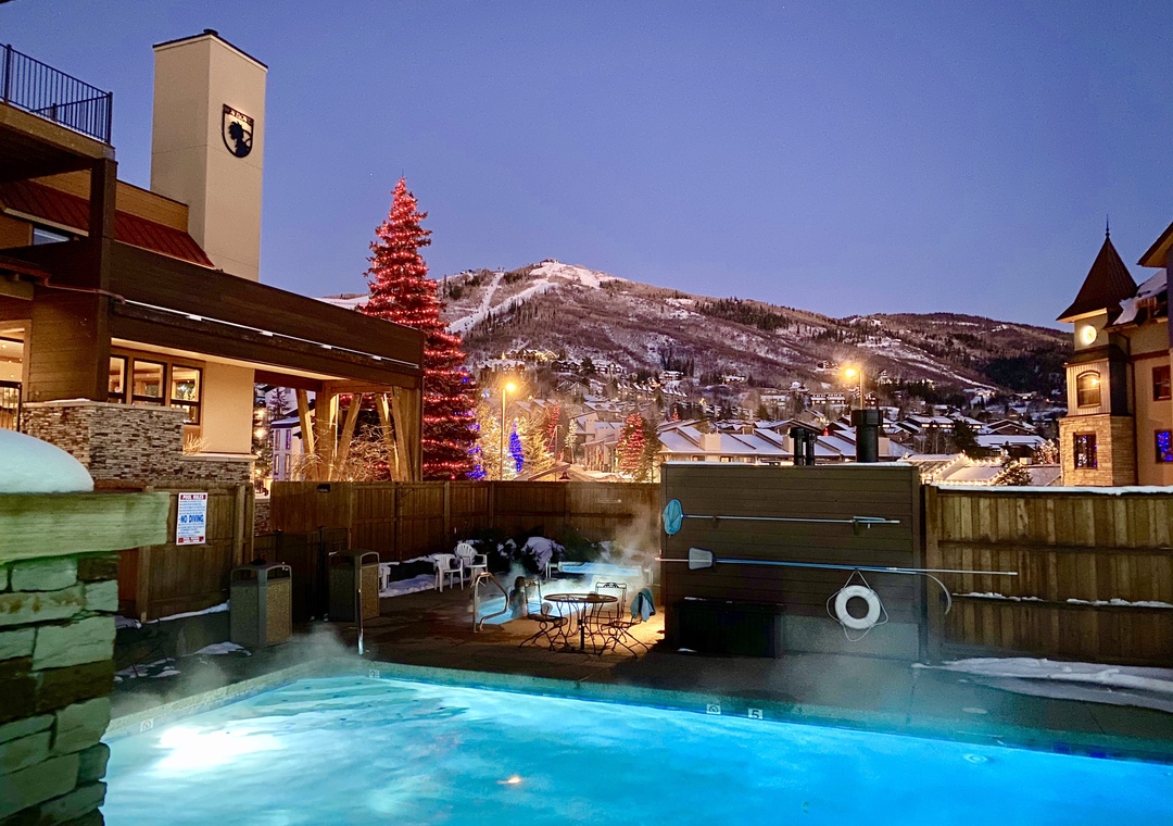 An outdoor pool and hot tub area glowing at twilight, framed by festive holiday lights and snow-dusted mountain slopes in the distance.