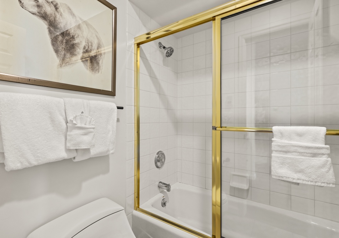 Your pristine bathroom features a glass-enclosed tub-shower combo with elegant gold accents and fresh white towels waiting for your arrival.