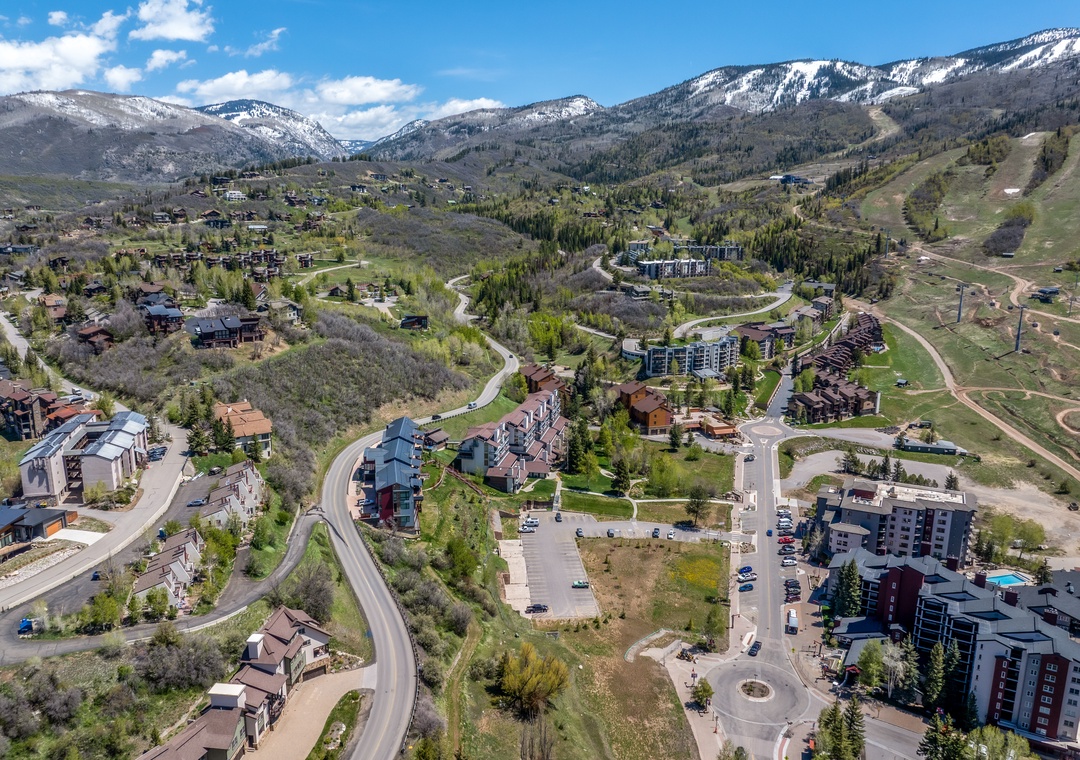 An aerial view reveals a mountain valley community nestled among evergreen forests, with snow-dusted peaks rising in the distance and winding roads connecting the residential neighborhood.