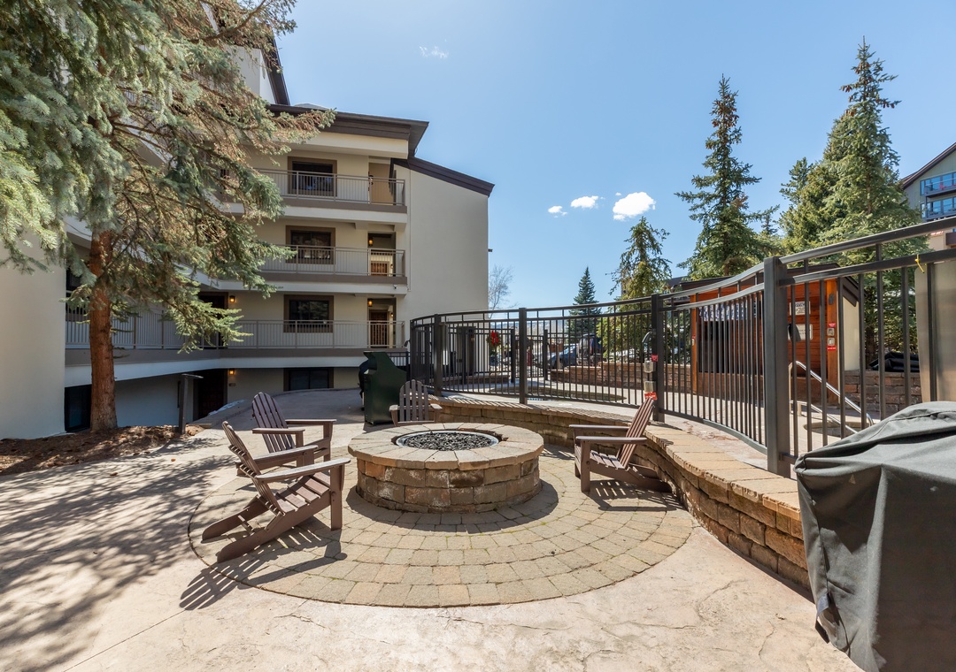 An outdoor stone courtyard with fire pit, Adirondack chairs, and BBQ area surrounded by evergreen trees—perfect for evening gatherings under open skies.