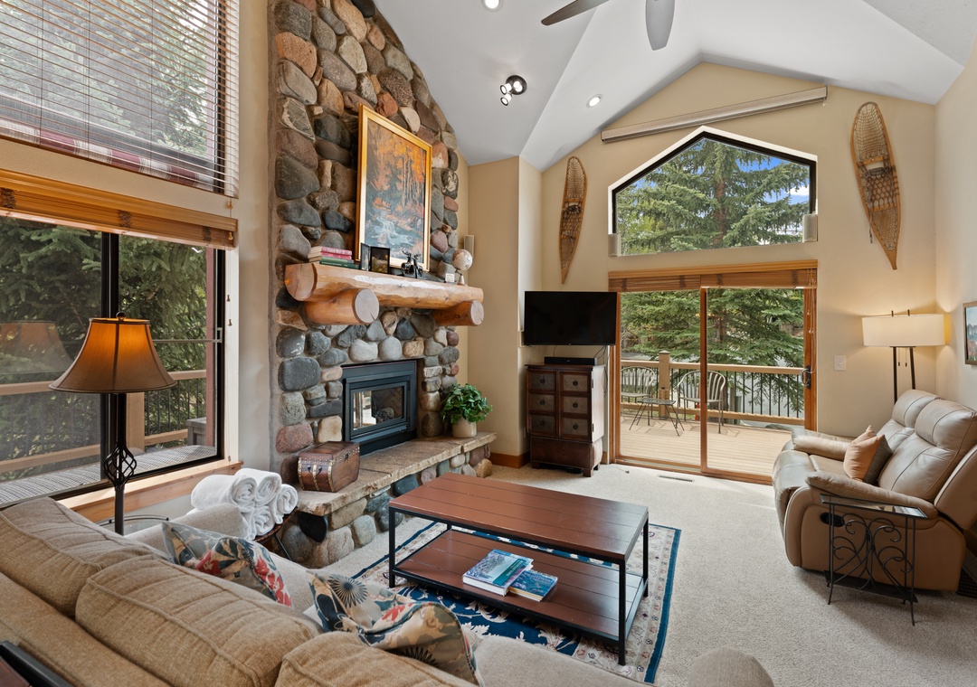 Relax in your mountain-retreat living room featuring soaring vaulted ceilings, a stunning stone fireplace, and private balcony access with forest views to complete your alpine escape.