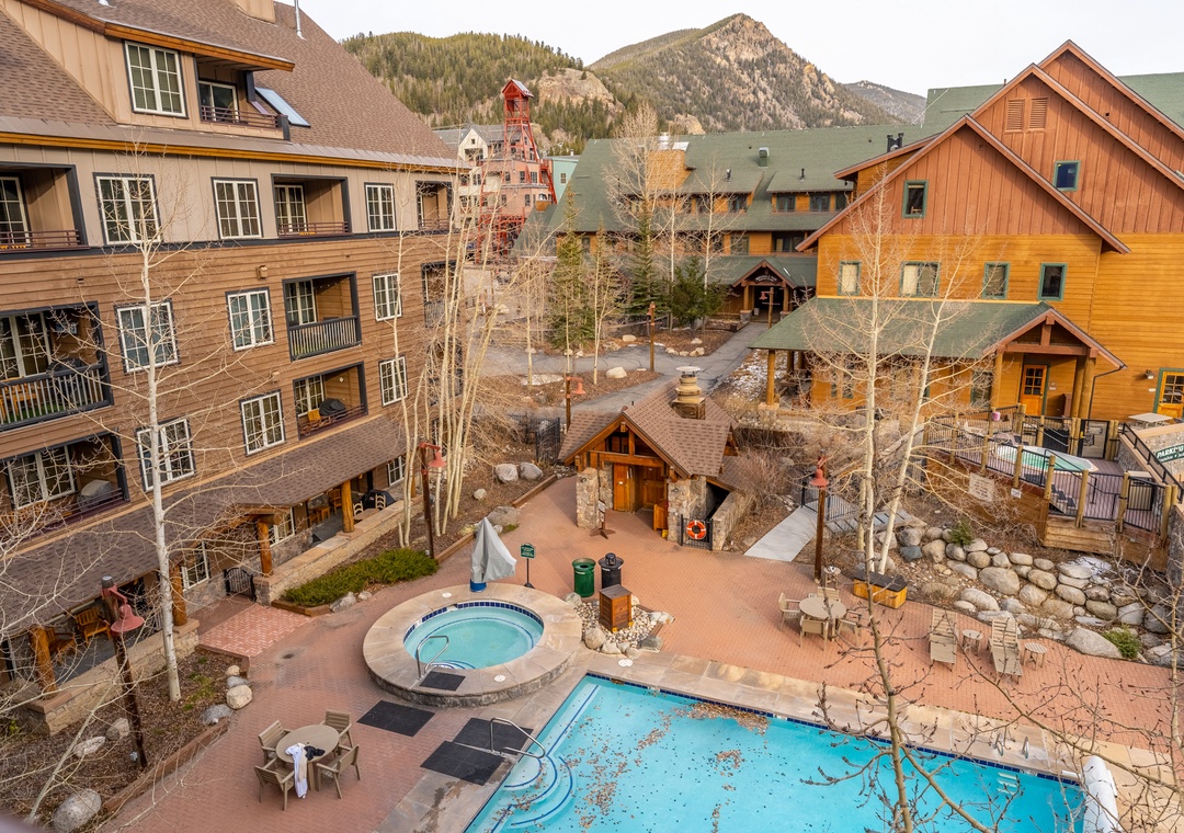 Mountain resort complex featuring rustic lodges, outdoor pools, and hot tub surrounded by dramatic alpine peaks and forested terrain.