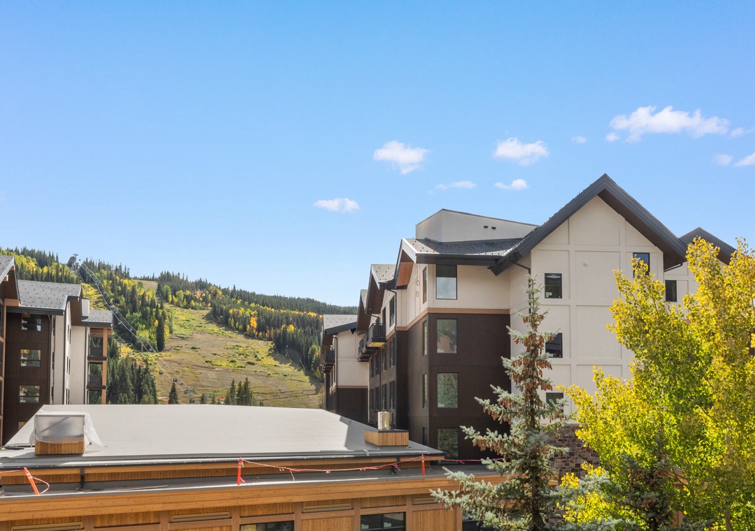 Modern mountain village buildings surrounded by golden autumn foliage and forest-covered slopes under bright blue skies.