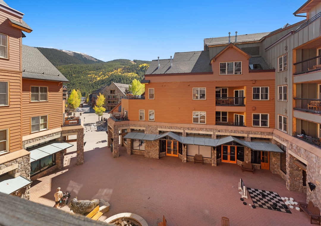 Mountain resort courtyard with stone and timber architecture, surrounded by forested peaks and autumn colors in the distance.