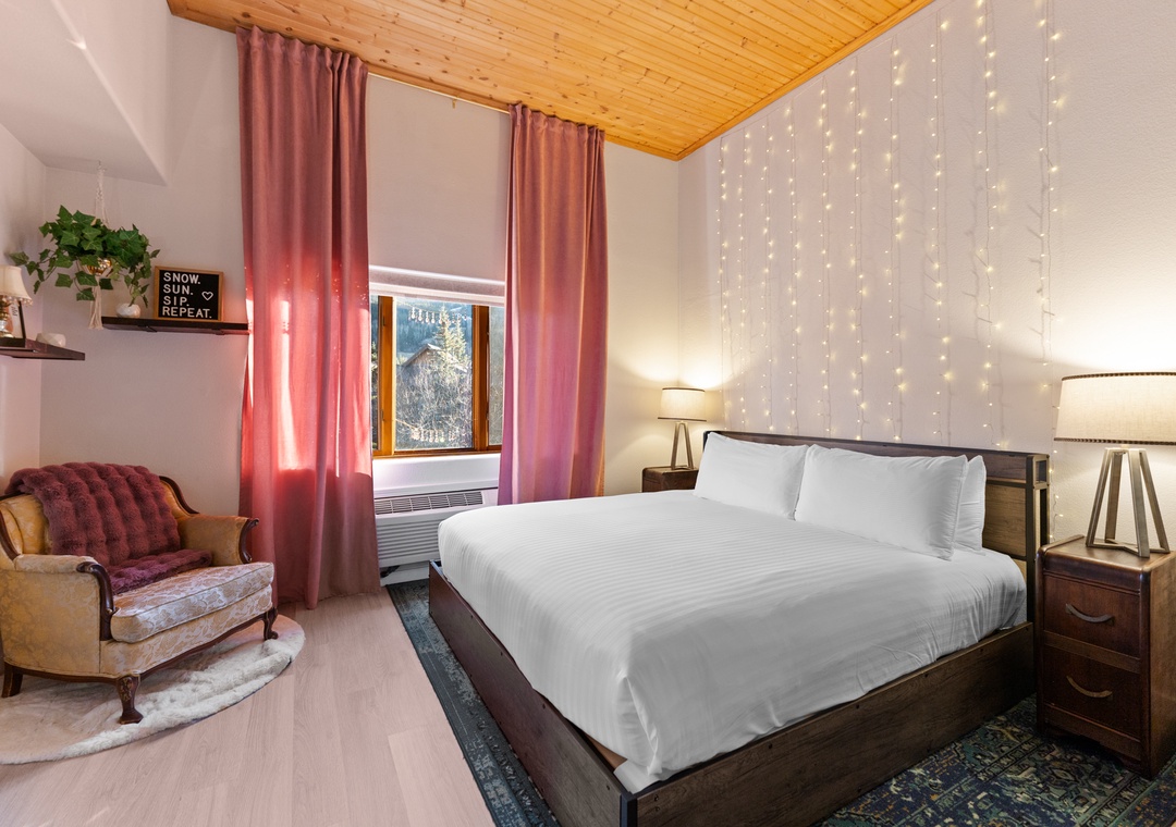 Sink into crisp white linens under twinkling fairy lights in your cozy mountain retreat with warm wood ceiling and vintage charm.