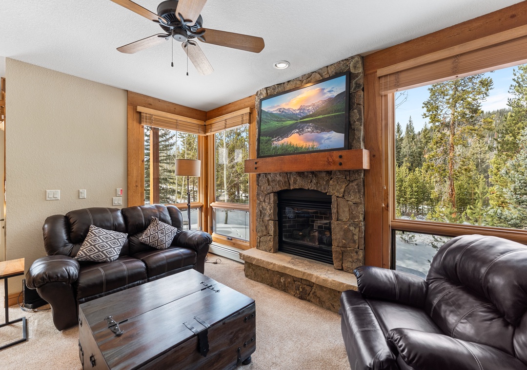 Cozy up by the stone fireplace in your living room, where leather seating and forest views create the perfect mountain retreat atmosphere.