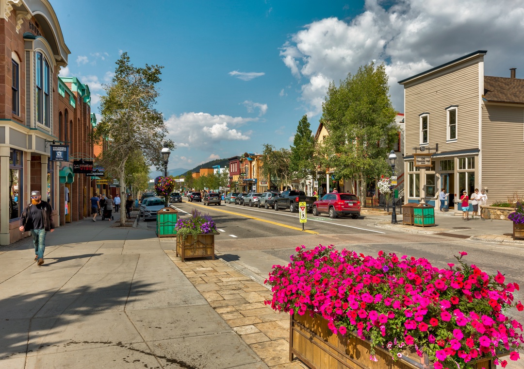 A charming historic downtown district with colorful storefronts and flower-lined streets. Mountains rise in the distance beneath summer skies, offering convenient access to local dining and shopping.