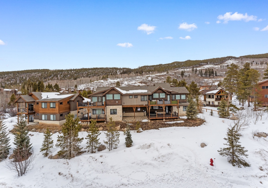 Modern mountain lodge nestled in pristine winter landscape with snow-covered peaks and forested hills in the background.