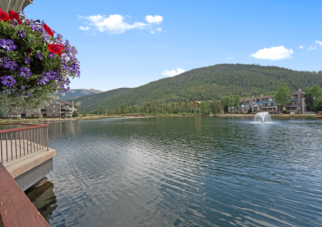 Serene mountain lake surrounded by forested hills and residential buildings, with a decorative fountain creating gentle ripples across the water.