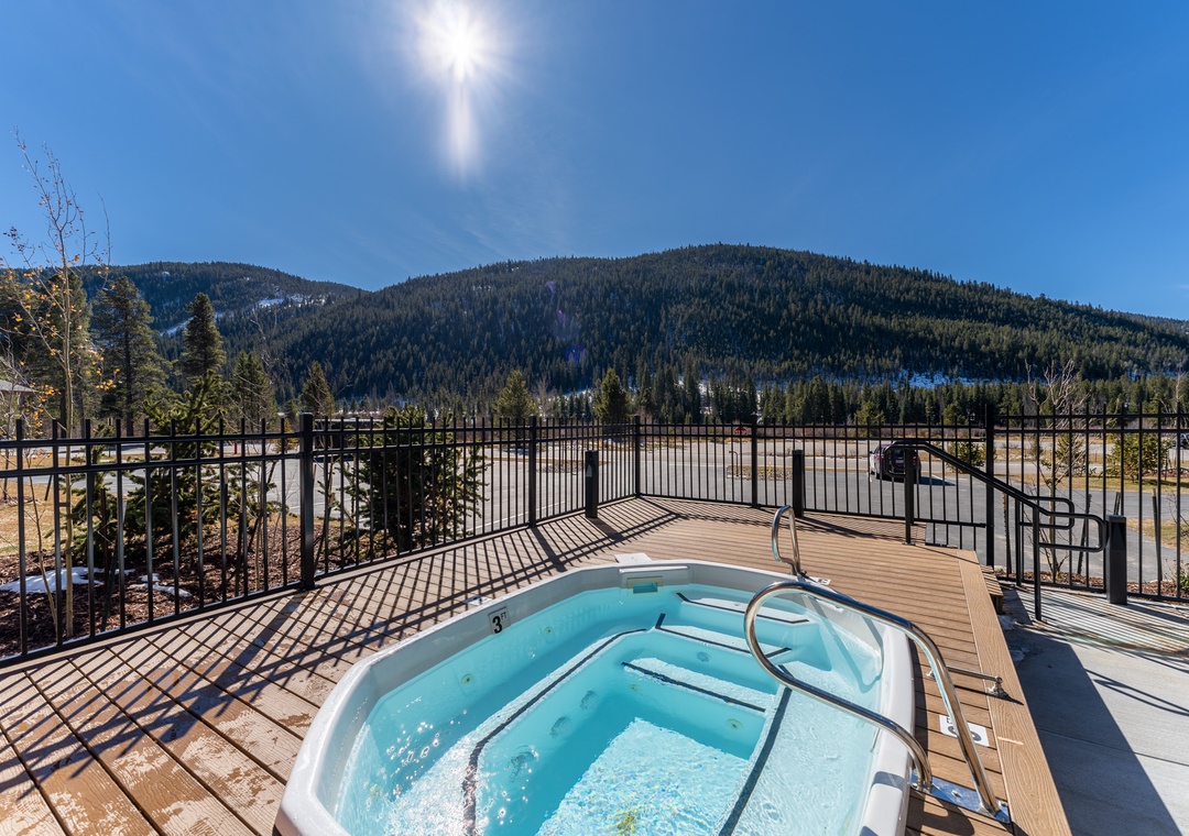 Hot tub on outdoor deck surrounded by snow-covered mountains and forest during a bright sunny winter day.