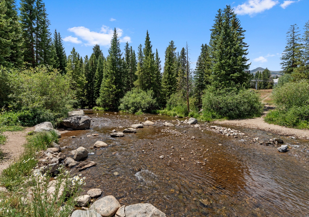 A peaceful creek winds through the property's natural landscape, framed by tall evergreens and mountain views—a perfect spot for morning walks or quiet reflection.