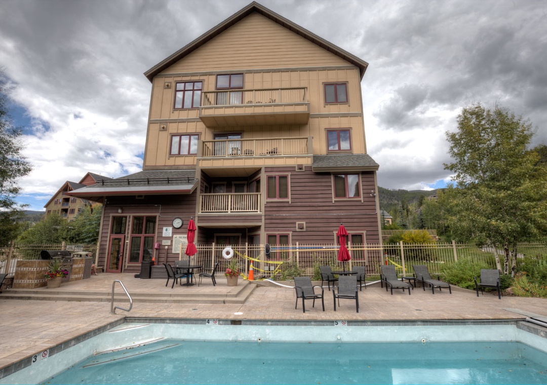 Multi-story mountain property featuring outdoor pool, patio seating, and scenic forest surroundings under dramatic cloudy skies.