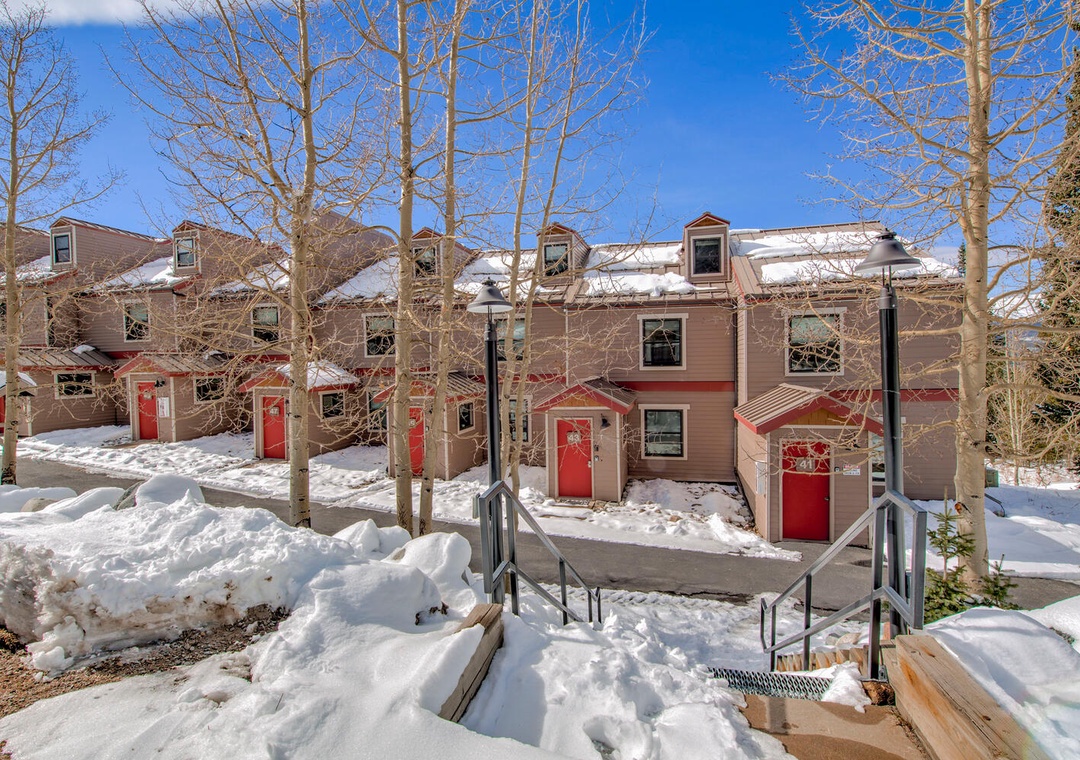 Charming winter wonderland awaits with snow-covered buildings and red doors creating a cozy mountain retreat atmosphere.