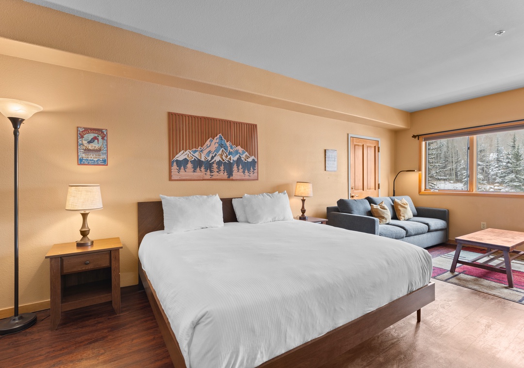 Relax in your cozy mountain retreat with a plush king bed, comfortable seating area, and serene winter views through large windows. Warm lighting and rustic touches create the perfect alpine escape.